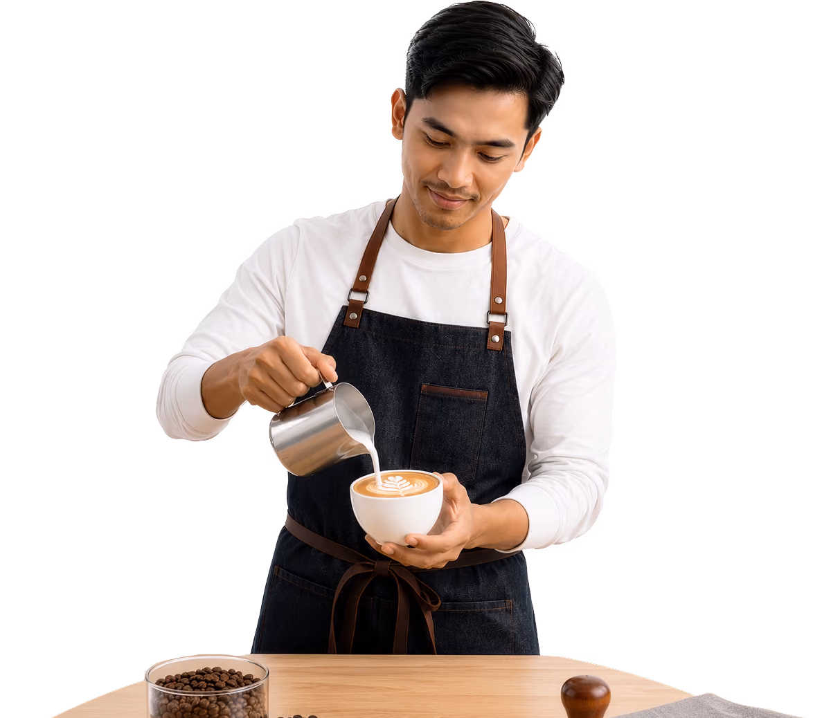 Barista in apron pouring milk into a cup of coffee with latte art on a wooden table.