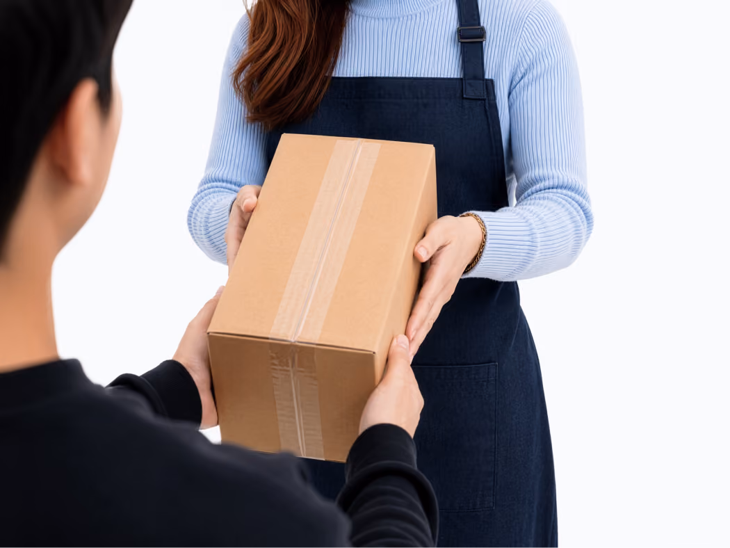 Woman in blue sweater and apron handing a taped cardboard box to a person in black.