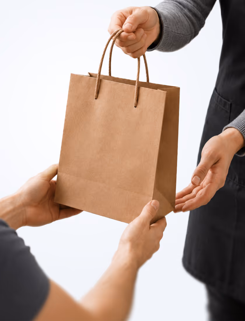 Person handing over a brown paper shopping bag to another person wearing a gray sleeve and black apron.